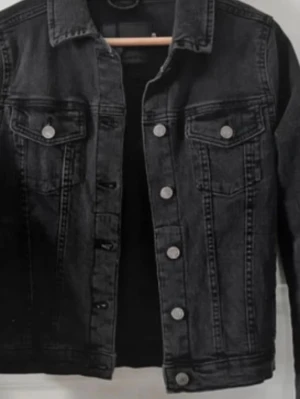 Svart klassisk jeansjacka - Snygg svart jeansjacka med silverfärgade knappar och två bröstfickor med lock. Jackan har en rak passform och klassisk krage. Perfekt att slänga över en hoodie eller t-shirt för en cool look.