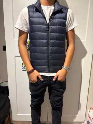 Uniqlo väst storlek xs  - Snygg mörkblå dunväst med quiltad design och hög krage. Västen har dragkedja framtill och är tillverkad i ett lätt material som ger bra värme utan att kännas klumpig. Perfekt att slänga över en hoodie eller t-shirt för en casual look.