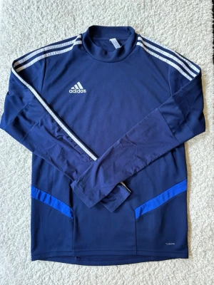 Mörkblå långärmad träningströja Adidas - Mörkblå långärmad träningströja från Adidas med vita ränder på axlarna och logga på bröstet. Tröjan har blå detaljer vid sidorna och är tillverkad i ett lätt, tekniskt material som andas. Perfekt för träning eller uppvärmning.