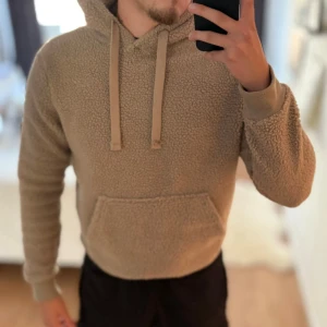Beige teddy hoodie med huva - Mysig beige teddy hoodie med huva och dragsnören. Hoodien har en stor magficka och ribbade muddar vid ärmsluten. Perfekt för dig som gillar en avslappnad och trendig stil. Materialet är fluffigt och ger en skön känsla.