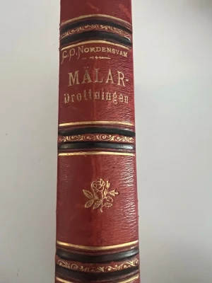 Mälardrottningen av G.P. Nordensvan - Klassisk roman med röd läderrygg och guldfolierad text samt dekorativa detaljer. Bokens rygg har en elegant ros i guld och vackra svarta och guldiga linjer. Perfekt för dig som gillar äldre skönlitteratur och snygga bokryggar i bokhyllan.
