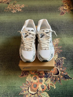 Vita Asics sneakers med mesh och silverdetaljer - Snygga vita Asics sneakers med meshpaneler och silverfärgade detaljer. Skorna har klassisk låg profil, vita snören och en robust sula med svart och grått mönster. Perfekta för dig som gillar sportig och clean stil.