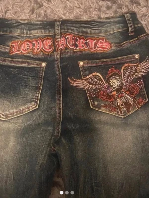 Love Hurts jeans med brodyr - Unika blå jeans med slitningar och broderad texten 'LOVE HURTS' bak samt en stor fickbrodyr med ängel och vingar. Låg midja och bootcut-modell, perfekta för dig som gillar statement-plagg och detaljer. Materialet är klassisk denim i bomull.