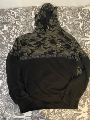 Philipp Plein hoodie med camomönster - Snygg hoodie från Philipp Plein med camomönstrad ovandel och svart nederdel. Huva med dragsko och metallogo på bröstet. Band med Philipp Plein-tryck går tvärs över bröstet. Perfekt för dig som gillar streetwear och vill sticka ut.