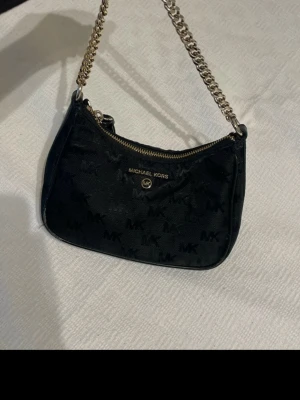 Svart axelremsväska Michael Kors - Snygg svart handväska från Michael Kors med guldfärgad kedjerem och dragkedja. Väskan har diskret MK-mönster över hela ytan och logga framtill. Perfekt storlek för det viktigaste och ger en lyxig touch till din outfit.