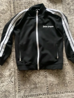 Svart track jacket från Palm Angels - Svart track jacket från Palm Angels med vita ränder längs ärmarna och vit dragkedja. Jackan har två fickor med dragkedja och logga på bröstet. Perfekt för en sportig och trendig look.