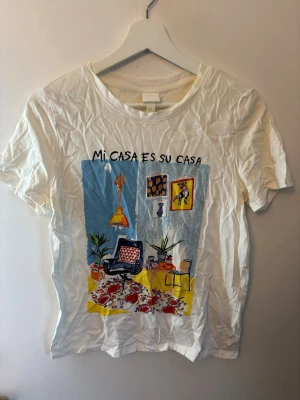 Vit t-shirt med färgglatt tryck - Vit t-shirt med korta ärmar och ett färgglatt tryck framtill med texten 'Mi casa es su casa'. Motivet visar ett inrett rum med detaljer i blått, gult, rött och svart. Rund halsringning och avslappnad passform. Perfekt för dig som gillar unika prints.
