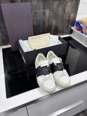 Valentino Garavani vita sneakers - Snygga vita sneakers från Valentino Garavani med bred svart läderdetalj över vristen. Klassisk låg modell med rund tå och vita snören. Kommer med originalkartong och dustbag. Perfekta för dig som vill ha en stilren och exklusiv look. Hör av dig vid minsta fundering om skorna!💯