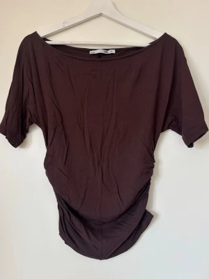 Brun loose t-shirt med bred hals - Säljer en brun t-shirt med bred, nästan offshoulder halsringning och loose passform. T-shirten har korta ärmar och är gjord i mjuk bomull, perfekt för en avslappnad stil. Enkel och stilren design utan mönster eller tryck.
