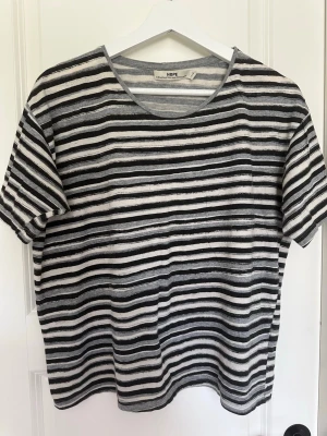 Randig t-shirt från HOPE - Säljer en randig t-shirt från HOPE i svart, grått och vitt. T-shirten har korta ärmar, rund halsringning och är gjord i mjuk bomull. Perfekt för en avslappnad och stilren look.