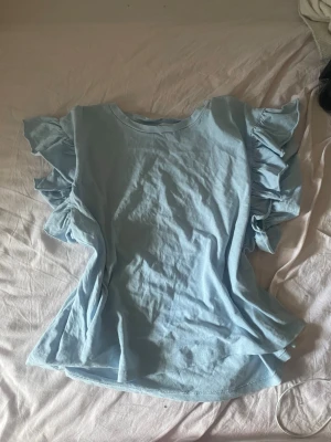 Ljusblå volangtopp från Zara - Supersöt ljusblå topp från Zara med volangdetaljer längs ärmhålen. Toppen är ärmlös och har en avslappnad passform, perfekt för dig som gillar en feminin och trendig stil. Tillverkad i mjuk bomull som känns skön mot huden.