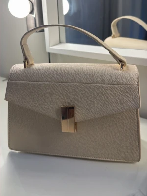 Beige handväska med guldiga detaljer - Stilren beige handväska i strukturmönstrat material med topphandtag och avtagbar axelrem. Väskan har ett elegant guldigt spänne framtill och en klassisk, rektangulär form. Perfekt accessoar för en trendig look.