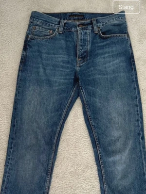 Blå jeans från Nudie Jeans - Säljer ett par klassiska blå jeans från Nudie Jeans med raka ben och fem fickor. Jeansen har en snygg tvättad look, kontrastsömmar och Nudie-loggan på bakfickan samt patch i läder bak i midjan. Perfekta till din vardagsstil! Storlek 28/30