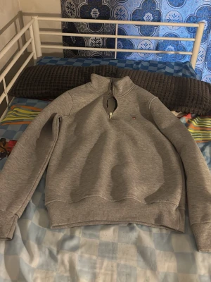 Grå halvzip sweatshirt från Gant - Snygg grå sweatshirt från Gant med halv dragkedja och liten broderad logga på bröstet. Tröjan har ribbade muddar vid ärmslut och nederkant samt en hög krage. Perfekt till chill eller skola.