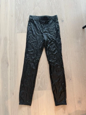 Svarta glansiga leggings - Säljer ett par svarta leggings med glansig finish och hög midja. De har en smal passform och är tillverkade i ett stretchigt material som sitter tajt längs benen. Perfekta för en trendig och bekväm look.