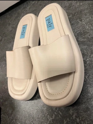 Beige chunky sandaler från Pair - Stilrena chunky sandaler från Pair i beige färg med bred rem över foten och fyrkantig tå. Tillverkade i ett slätt konstläder med vadderad innersula för extra komfort. Sulan är svart undertill och har räfflad struktur för bättre grepp. Ord 2100kr 
