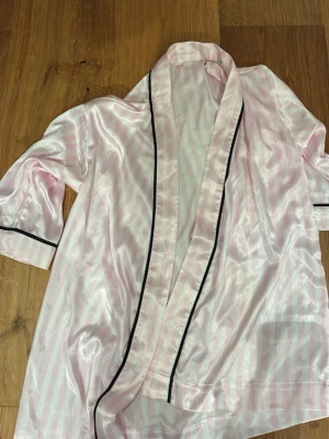 Rosa randig morgonrock Gina Tricot - Sidenliknande morgonrock från Gina Tricot i ljusrosa och vita ränder med svarta kanter. Lätt och glansigt material, trekvartsärmar och öppen framtill. Perfekt för chilliga morgnar eller kvällar hemma.