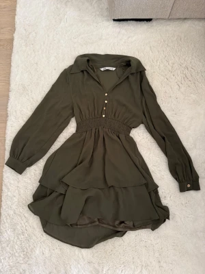 Olivgrön kort klänning med volanger - Snygg olivgrön kortklänning med långa ärmar, volangdetaljer och markerad midja. Klänningen har krage, knappar framtill och är tillverkad i ett lätt, flowy material som ger en avslappnad men chic look.