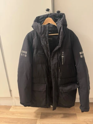 Svart dunjacka från Sail Racing - Svart dunjacka från Sail Racing med huva och dragkedja framtill. Jackan har två stora fickor, logga på bröstet och båda ärmarna samt en sportig design. Perfekt för kalla vinterdagar och håller dig varm med sin vaddering.