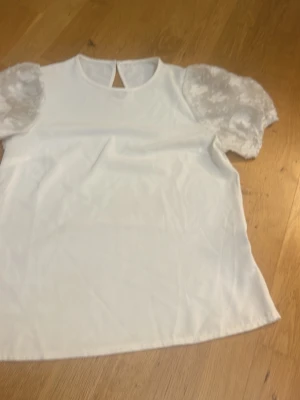 Vit topp med puffärmar i spets - Snygg vit topp med genomskinliga puffärmar i spets och rund halsringning. Toppen har en liten knapp i nacken och är i en rak modell. Perfekt för dig som vill ha en söt och trendig look.