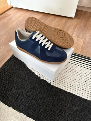 Marinblå maison margiellas  - Snygga marinblå sneakers med vita skosnören och kontrasterande ljusbrun gummisula. Ovandelen är i en mix av mocka och slätare material för en cool look. Perfekta för dig som vill ha stilrena och trendiga skor till vardagsoutfiten.