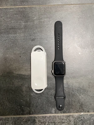 Apple Watch  - Snygg svart smartklocka med rektangulär skärm och matchande svart silikonarmband. Klockan har en metallram och kommer med en vit laddare. Perfekt för dig som vill ha koll på notiser och hälsa direkt på handleden.