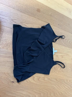 Svart omlott linnetopp med volang - Svart omlott linnetopp med smala axelband och volangdetalj framtill. Toppen har en croppad passform och är perfekt för en trendig look. Tillverkad i mjukt material som känns skönt mot huden.