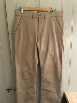 Beige chinos med rak passform - Snygga beige chinos med rak passform och klassisk design. Byxorna har diskreta sidfickor och är tillverkade i ett mjukt bomullsmaterial som känns bekvämt hela dagen. Perfekta för en stilren och avslappnad look.