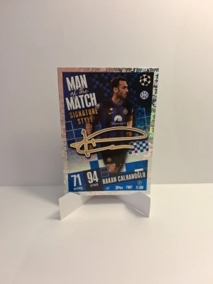 Match Attax Man of the Match-kort - Säljer ett Match Attax samlarkort med Hakan Calhanoglu från Inter. Kortet har texten 'Man of the Match Signature Style' och är dekorerat med blå och guldiga detaljer samt en tryckt autograf. Perfekt för dig som gillar fotboll och samlarkort.