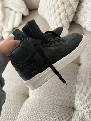 Svarta high top sneakers från Puma - Snygga svarta high top sneakers från Puma med vit sula. Skorna har klassisk snörning, perforerad tå och diskret Puma-logga på sidan och plösen. Tillverkade i syntetmaterial med sportig vibe och tidlös design. Fodrade underbara sneakers för kallare dagar 