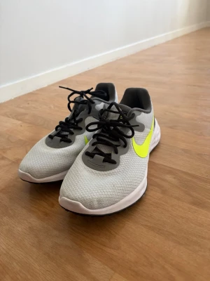 Grå Nike sneakers med neongul swoosh - Snygga sneakers från Nike i grå mesh med svart hälparti och svart snörning. Sidan pryds av en stor neongul swoosh som ger skon en sportig vibe. Sulan är vit och ger bra komfort. Perfekta för dig som gillar stilrena men ändå färgglada detaljer.