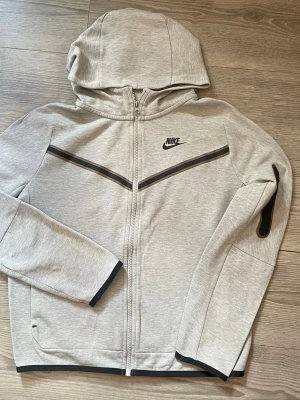 Nike tech fleece  - Snygg och väldeligt skön Nike tech från Nike med dragkedja framtill och svart Nike-logga på bröstet. Hoodien har svarta detaljer längs bröstet och ärmslut samt en praktisk huva. Tillverkad i mjukt sweatshirtmaterial, perfekt för en avslappnad streetwear-look.