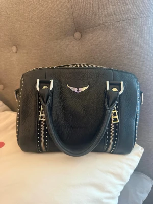 Zadig & Voltaire  Sunny bag - Cool svart handväska i skinnimitation med silverfärgade detaljer, dubbla handtag och dekorativa dragkedjor framtill. Väskan har små nitar längs sidorna och ett litet emblem i silver och lila på framsidan. Köptes nypris för 5100kr på Jackie. Använd sparsamt. Priset går ABSOLUT att diskutera!!! 💕💕