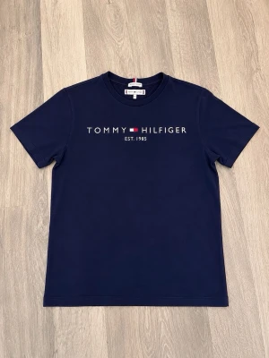 Marinblå t-shirt från Tommy Hilfiger - Snygg marinblå t-shirt från Tommy Hilfiger med klassiskt tryck på bröstet och rund halsringning. T-shirten har korta ärmar och är tillverkad i mjuk bomull, perfekt för en avslappnad och stilren look.