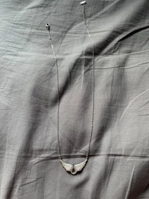 Silverfärgat halsband med vingar - Snyggt halsband i silverfärg med ett hängsmycke format som vingar, täckta av glittrande stenar. Kedjan är tunn och smidig, och vingarna ger ett coolt och unikt intryck. Perfekt för dig som gillar detaljer som sticker ut.