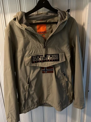 Beige vindjacka från Napapijri - Snygg beige vindjacka från Napapijri med stor logga och norsk flagga på bröstet. Jackan har huva, halv dragkedja framtill, två sidofickor med dragkedja och meshfoder. Perfekt för blåsiga dagar, passar för S och M