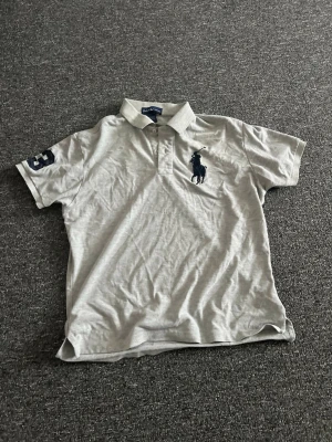 Ljusgrå pikétröja från Ralph Laure n - Klassisk ljusgrå pikétröja från Ralph Laure n med korta ärmar och krage. Stor mörkblå logga på bröstet och siffran 3 på ena ärmen. Perfekt till jeans eller shorts för en clean och sportig look.
