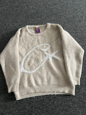 Beige stickad tröja med vit detalj - Mysig beige stickad tröja med en stor vit bokstav på framsidan. Tröjan har rund halsringning, ribbade muddar och avslut. Perfekt för dig som gillar en avslappnad och trendig stil. Materialet känns mjukt och skönt mot huden.