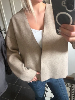 Beige oversized kofta med v-ringning - Säljer en beige oversized kofta med djup v-ringning och raka, vida ärmar. Koftan är enkel och stilren utan synliga knappar eller detaljer, perfekt att slänga över en topp. Materialet ser mjukt och lite tjockare ut, vilket gör den skön att bära.