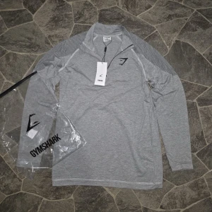 Grå långärmad träningströja Gymshark - Snygg grå långärmad träningströja från Gymshark med halv dragkedja framtill och diskret logga på bröstet. Tröjan har meshdetaljer på axlarna för extra ventilation och är tillverkad i ett mjukt, stretchigt material som passar perfekt till gymmet.