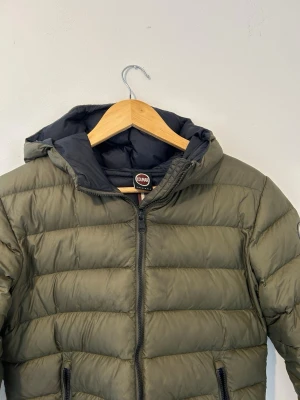Olivgrön dunjacka från Colmar - Snygg olivgrön dunjacka från Colmar med huva och dragkedja framtill. Jackan har två fickor med dragkedja och Colmar-logga på ärmen. Perfekt för kalla dagar och har en klassisk, puffig design.