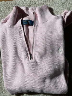 Ljusrosa halvzip från Polo Ralph Lauren - Ljusrosa tröja med halv dragkedja från Polo Ralph Lauren. Klassisk design med ribbade muddar och broderad logga på bröstet. Perfekt för en avslappnad men ändå stilren look. Tillverkad i mjukt bomullsmaterial.
