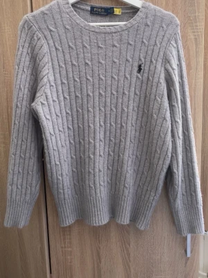 Grå kabelstickad tröja Polo Ralph Lauren - Snygg grå kabelstickad tröja från Polo Ralph Lauren med rund halsringning och broderad logga på bröstet. Tröjan har långa ärmar och ribbstickade muddar vid ärmslut och nederkant. Perfekt för lager-på-lager-stil.