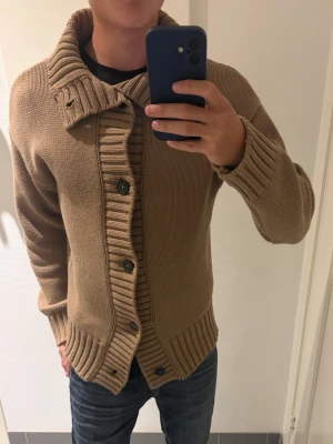 Beige stickad kofta med krage - Säljer en beige stickad kofta med stora svarta knappar och bred ribbad kant. Koftan har en hög, ribbad krage och långa ärmar med ribbade muddar. Bra skick, skriv gärna vid intresse!