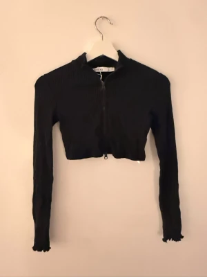Svart croppad ribbad polotopp - Snygg svart croppad polotopp med långa ärmar och ribbad struktur. Toppen har dragkedja framtill och en tight passform som framhäver figuren. Perfekt att styla med höga jeans eller kjol.