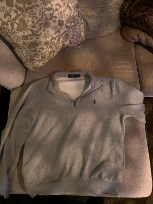 Grå halvzip tröja från Polo Ralph Lauren - Snygg grå halvzip tröja från Polo Ralph Lauren med klassisk broderad logga på bröstet. Tröjan har hög krage och ribbade muddar vid ärmslut och nederkant. Perfekt för lager på lager och enkel att matcha med jeans eller chinos.