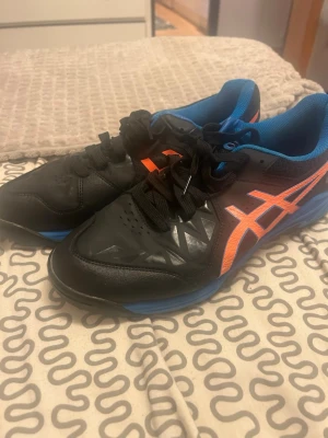 Svarta Asics Gel-Peake sportskor - Säljer ett par svarta Asics Gel-Peake sportskor med blå sula och orangea detaljer. Skorna har klassisk snörning, låg profil och är tillverkade i slitstarkt syntetmaterial med ventilerande mesh på insidan. Perfekta för inomhus- eller utomhusträning.