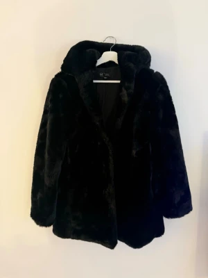 Svart fuskpälsjacka med huva Zara - Säljer en svart fuskpälsjacka med huva. Jackan har en mjuk och fluffig känsla, rak passform och långa ärmar. Perfekt för kalla dagar när du vill hålla dig varm och samtidigt se snygg ut. Stängs framtill och har en stilren look.