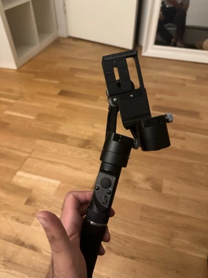 Svart kamerastabiliseringsgimbal - Säljer en svart kamerastabiliseringsgimbal med flera justerbara leder och kontrollknappar på handtaget. Perfekt för att få stabila videoklipp med din kamera eller mobil. Tillverkad i metall och plast med en stilren design.