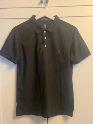 Svart pikétröja från Polo Ralph Lauren - Klassisk svart pikétröja från Polo Ralph Lauren med korta ärmar och tre vita knappar framtill. Tröjan har den ikoniska röda logotypen broderad på bröstet och är tillverkad i bomull för en bekväm känsla.